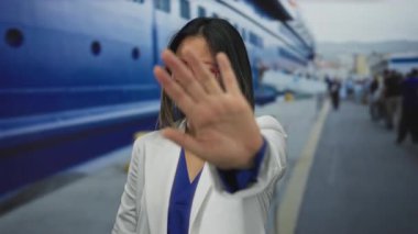 Beyaz ceketli genç Çinli bir kadın arka planda büyük bir tekneyle sahil limanında duraklama hareketi yapıyor. Seyahat ve denizcilik aktivitelerini çağrıştırıyor..