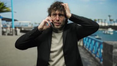 Takım elbiseli İspanyol bir adam deniz kenarında arka planda teknelerle telefon görüşmesi yaparken stresi dile getiriyor..