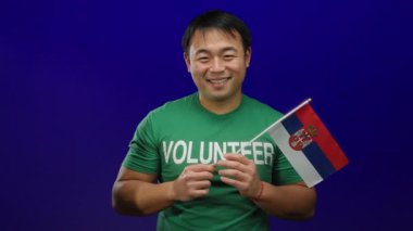 Sırbistanlı bayrak tutan gülümseyen genç adam mavi arka planda gönüllü tişört giyiyor.