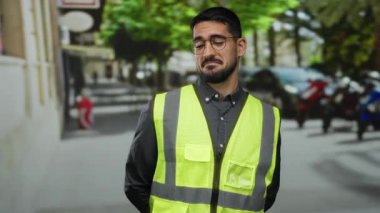 Yansıtıcı yelek giyen bir adam, şehrin arka planının bulanık olduğu bir sokakta düşünceli bir ifadeyle duruyor..