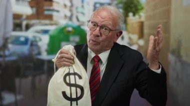 Takım elbiseli kıdemli bir adam açık hava restoranındaki terasta bir dolar işaretli bir çanta tutuyor. Kalabalık bir caddede para ve yolsuzluk temaları öneriyor..