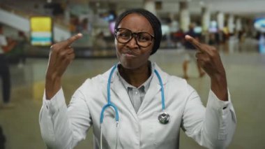 Laboratuvar önlüğü giymiş bir kadın havaalanı terminalinde elleri hareketli komik hareketler yapıyor. Etrafı gezginlerle çevrili mizah anlayışı gösteriyor..