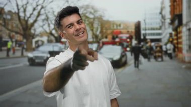 Beyaz tişörtlü genç adam canlı bir şehir caddesinde insanların yürüdüğü ve trafiğin geçtiği hareketli şehir atmosferini yükselterek gülümsüyor..