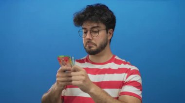 Sakallı genç adam, parlak mavi bir arkaplan karşısında İsviçre franklarını teftiş ediyor, çizgili bir gömlek ve gözlük takıyor, odaklanma ve tefekkür ediyor..