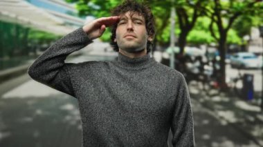 Koyu renk saçlı, gri kazaklı genç adam şehir caddesinde duruyor ve selam veriyor.