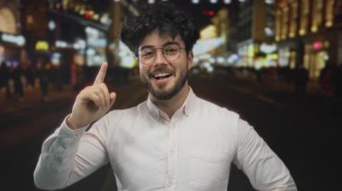 Gözlüklü, sakallı, şehir ışıklarıyla aydınlatılmış genç bir İspanyol gece açık havayı işaret ediyor. Bu da kalabalık bir şehir ortamında keşfedildiğini gösteriyor..