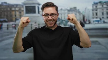 Gözlüklü genç adam şehir meydanında coşkuyla gülümsüyor. Arka planda tarihi bir mimari var. Olumlu bir ruh halini gösteriyor..