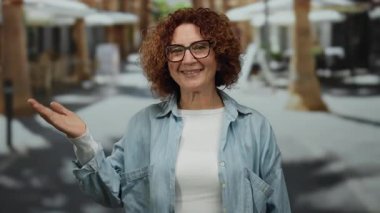 Güneşli bir şehir caddesinde gülümseyen, gözlüklü ve kot gömlekli neşeli bir ifadeyle, açık havada canlı bir şehir atmosferi yakalıyor..