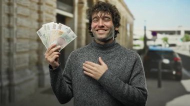 Elleri göğsünde gülümseyen İspanyol bir adam, şehir sokaklarında açık havada Czech Koruna banknotlarını tutuyor ve mutluluğu ve finansal başarıyı gösteriyor..