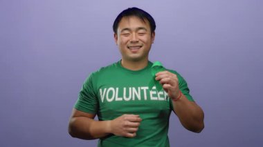 Yeşil gönüllü gömleği giyen genç adam mor arka planda kurdele tutuyor. Kameraya gülümsüyor, yardım ve farkındalık taşıyor..