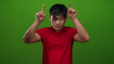 Kırmızı tişörtlü genç Çinli adam yeşil bir arka plan duvarına boynuzlarını şakaya vurarak neşeli ve genç bir hava veriyor..