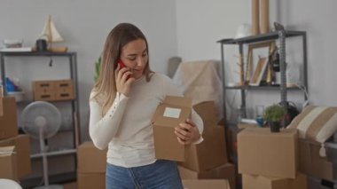 Yeni bir evde kutu ve raflarla çevrili bir evde telefonla konuşan bir kadın, taşınmayı ya da yeni bir eve yerleşmeyi ima ediyor..