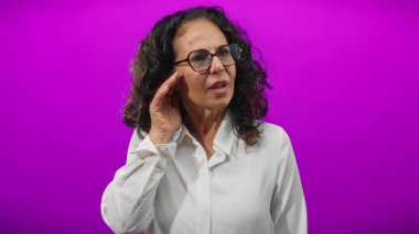 Kadın, canlı bir mor arka planda gülümseyen jestler dinler. Kulağında açık beyaz gömlek ve gözlük takan etkileyici bir elle merak ve nişanı somutlaştırır..