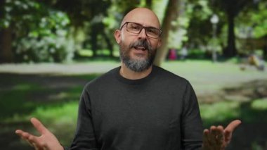 Gözlüklü, sakallı ve dövmeli bir adam güneşli bir park ortamında gülümsüyor, yemyeşil ortamda rahat ve kendinden emin bir tavır sergiliyor..