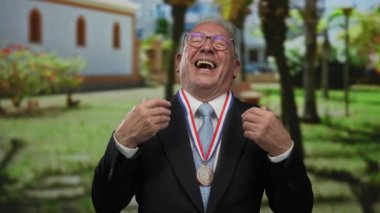 Takım elbiseli, güneş parkında neşeyle gülümseyen, neşeli tavırlarıyla başarısını sergileyen gözlüklü ve madalyalı bir adam..