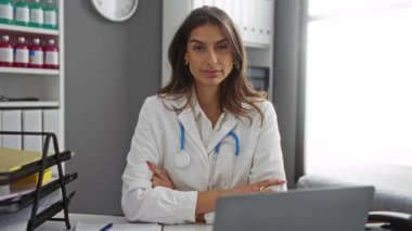 Klinikte kolları bağlı, steteskop takan kendine güvenen bir kadın doktor. Tıp bürosunda profesyonellik ve yakınlık gösteriyor..