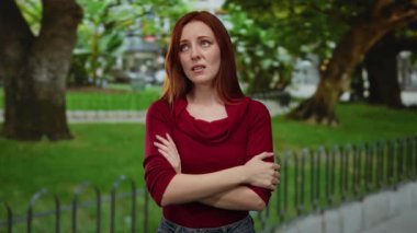 Kızıl saçlı bir kadın, kollarını kavuşturmuş bir şekilde, yemyeşil bir parkın içinde üzgün bir şekilde duruyor ve açık havada sakin bir ortam yaratıyor..