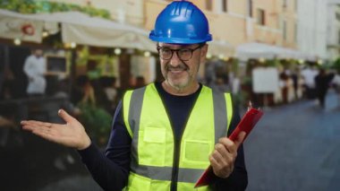 Şantiye miğferi ve yelek giymiş, kentin hareketli caddesinde elinde bir pano tutan, kendine güvenen gülümsemesini sergileyen olgun İspanyol bir adam..