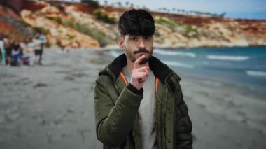 Genç İspanyol bir adam, sakin bir plaj ve okyanus arkaplanı ile düşünceli bir şekilde deniz kenarında el kol hareketi yapıyor, tefekkür ediyor ve bir dış mekâna odaklanıyor..