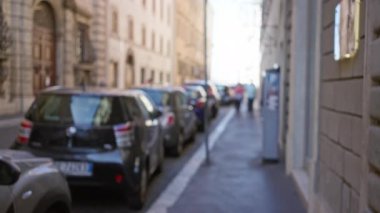 Odaklanamayan görüntü, Roma caddesinin ikonik görüntüsünü çekiyor. Bulanık park halindeki arabalarla, kaldırımlı yolda, İtalyan kentsel yaşam tarzının özünü yakalayan tarihi cephelerle dolu..