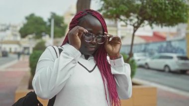 Gözlüklü bir kadın, şehir caddesindeki bir sokakta gözlerini ayarlıyor. Bulutlu gökyüzünün altında canlı pembe örgüler sergiliyor. Arka planda park etmiş arabalar ve binalar var..