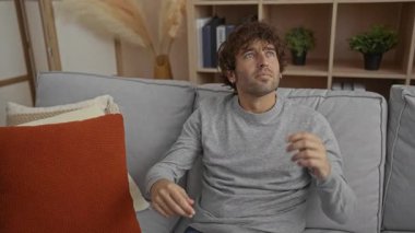 Gri kazaklı, kıvırcık saçlı genç bir adam, rahat kanepe ve dekoratif yastıklarla dolu bir oturma odasında el kol hareketi yapıyor, modern ev dekorasyonu elementleriyle çevrili, rahat bir dairede..