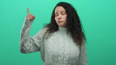 Süveter giyen genç İspanyol kadın, yeşil duvar arkaplanını işaret ediyor. Onaylamadığını belirtiyor ve yön gösteriyor..
