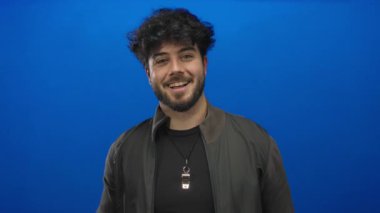 Sakallı, parlak mavi arka planda dikilen, rahat ve kendinden emin bir tavır takınan, siyah ceketli, Latin asıllı genç bir adam..