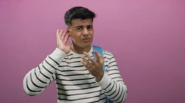 Genç adam pembe arka planda kafa karışıklığı gösteriyor, çizgili kazak ve sırt çantası giyiyor, merakını dile getiriyor ve dinliyor..