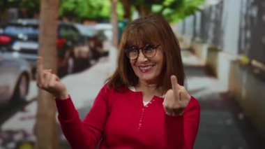 Kırmızı kazaklı İspanyol bir kadın şehir arka planında orta parmaklarıyla bir jest yapıyor, duygularını ve davranışlarını açık havada ifade ediyor..