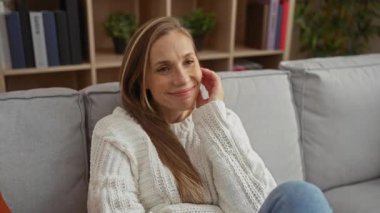 Oturma odasındaki rahat koltukta rahat bir şekilde gülümseyen bir kadın. Etrafı kitaplarla ve bitkilerle çevrili beyaz bir süveter giyiyordu. Evde huzurlu bir evin keyfini çıkarıyordu..