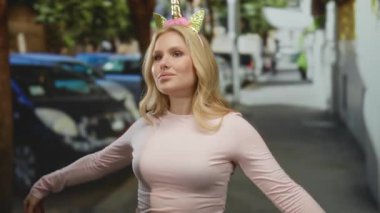 Sarışın kadın, tek boynuzlu at diademi ile neşeyle poz veriyor şehir caddesinde, fantezi ve eğlenceyi harmanlıyor, neşeli şehir havasını somutlaştırıyor.