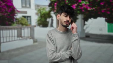 Siyah saçlı ve gri kazaklı genç adam arka planda çiçekleri bulanık bir şekilde şehir sokağında akıllı telefondan konuşuyor.