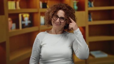 Kütüphanede gülümseyen, gözlük takan ve kafasını işaret eden bir kadın kitaplıklar arasında düşünce ve zeka gösterisi yapıyor..