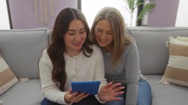 Anne ve kızı sıcak bir oturma odasında birlikte oturuyorlar, bir tablete bakarken iletişim ve sohbet anlarını paylaşıyorlar, aile sevgisini ve kapalı alan sıcaklığını gösteriyorlar..