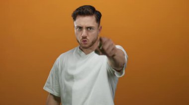 Sakallı genç adam, izole edilmiş turuncu arka planda kameraya kızgın bir şekilde el kol hareketi yapıyor. Günlük beyaz gömlek giyiyor..