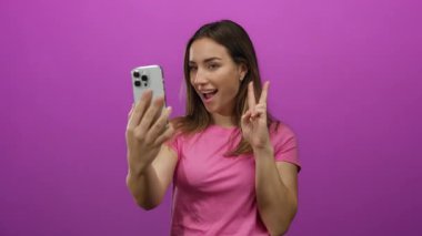 Pembe arka planda akıllı telefonlu selfie çeken bir kadın barış işareti yapıyor. Sarışın, izole edilmiş, mutlu, sıradan teknoloji modern portre..