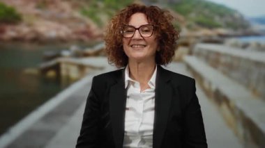 İspanyol kadın deniz kenarında gezinirken parmaklarıyla kalp hareketi yapıyor, siyah bir takım elbise ve gözlük takıyor..