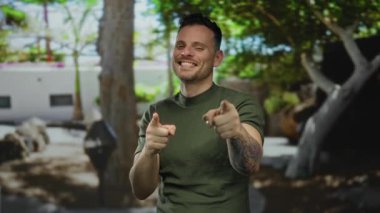 Yeşil gömlekli genç adam yemyeşil bir açık hava parkında poz verirken arka planda ağaçlarla birlikte, kendine güvenerek gülümseyerek dövmelerini sergiliyor..