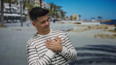 Plajda çizgili tişörtlü genç adam gözleri kapalı, elleri göğsünde, açık okyanusun kenarında, kıyı manzarası ve palmiye ağaçları arasında memnuniyetini ifade ediyor.