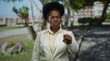 Park ortamında duran, beyaz ceketli, gözleri kapalı, ifadesiz, sakin, afro saç stili, ağaçlarla çevrili, gündüz sahnesi, doğal arka plan..