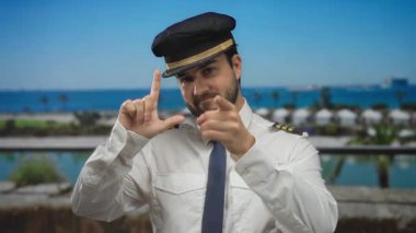 Pilot üniformalı İspanyol sakallı adam arka planda sakin mavi deniz ve palmiye ağaçlarıyla deniz kenarında ezik jestler yapıyor..