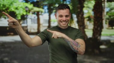 Dövmeleri olan genç bir adam gülümsüyor ve yemyeşil bir parkı işaret ediyor. Açık havada gündelik bir anı yakalıyor..