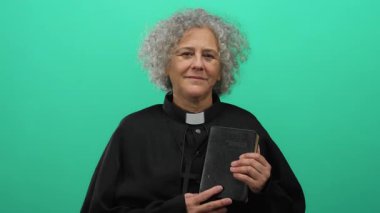 Gri saçlı bir rahip gibi giyinmiş bir kadın, izole edilmiş yeşil bir zemine karşı İncil 'i tutuyor, kameraya sıcak bir şekilde gülümsüyor, inanç ve maneviyatı somutlaştırıyor..