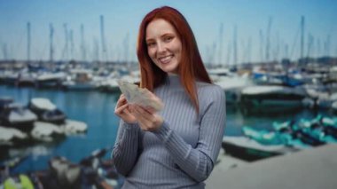 Elinde İsviçre banknotlarıyla gülümseyen bir kadın güneşli bir günde arka planda tekneler olan deniz manzaralı bir marinada.