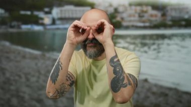 Sakallı ve dövmeli bir adam deniz kenarındaki el dürbününden bakıyor kumlu sahilde sıradan sarı bir gömlek giyiyor ve bulanık deniz altyapısı ile gülümsüyor.