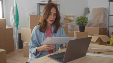 Dizüstü bilgisayarda çalışan bir kadın yeni dairede kutuları taşırken, belgeleri incelerken, masada yeşil bir bitki, günlük kıyafetler giyerken, rahat bir çalışma alanı yaratırken,.