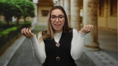 Gözlüklü genç bir kadın, arka planda tarihi mimari ve yeşillik ile çevrili düşünceli bir ifadeyle eski bir üniversitede el kol hareketi yapıyor.