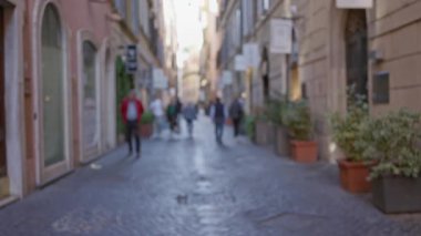 Roma 'daki sokak sahnesinde bulanık insanlar Bokeh etkisinden süzülen parke taşları, canlı vitrinleri ve güneş ışığıyla eski bir italyan caddesinde yürüyor..
