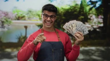 Açık hava parkında elinde Amerikan doları banknotları tutan güneş gibi doğal bir ortamda güvenle gülümseyen ve kırmızı süveter giyen bir adam..